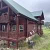 A rustic log cabin with a green metal roof nestles on the grassy hillside of Larimer County, CO. The cabin features multiple balconies, visible satellite dishes, and a stone foundation. Overcast skies hover above, while distant trees frame this picturesque Colorado scene.
