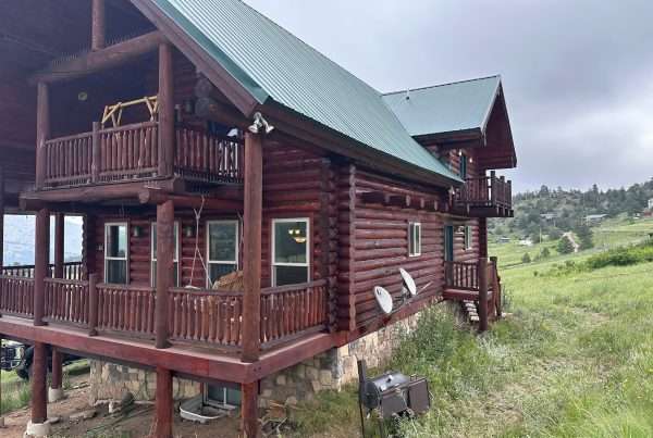 A rustic log cabin with a green metal roof nestles on the grassy hillside of Larimer County, CO. The cabin features multiple balconies, visible satellite dishes, and a stone foundation. Overcast skies hover above, while distant trees frame this picturesque Colorado scene.