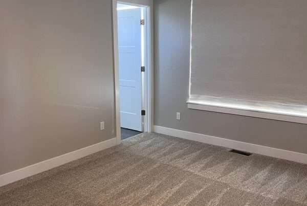 An empty room with beige carpet and gray walls, featuring a modern black ceiling fan. A doorway leads to another room, and a window with closed beige blinds is present. White baseboards offer contrast, enhancing the sleek look perfect for interior painting. Ideal for spaces in Larimer County, CO.