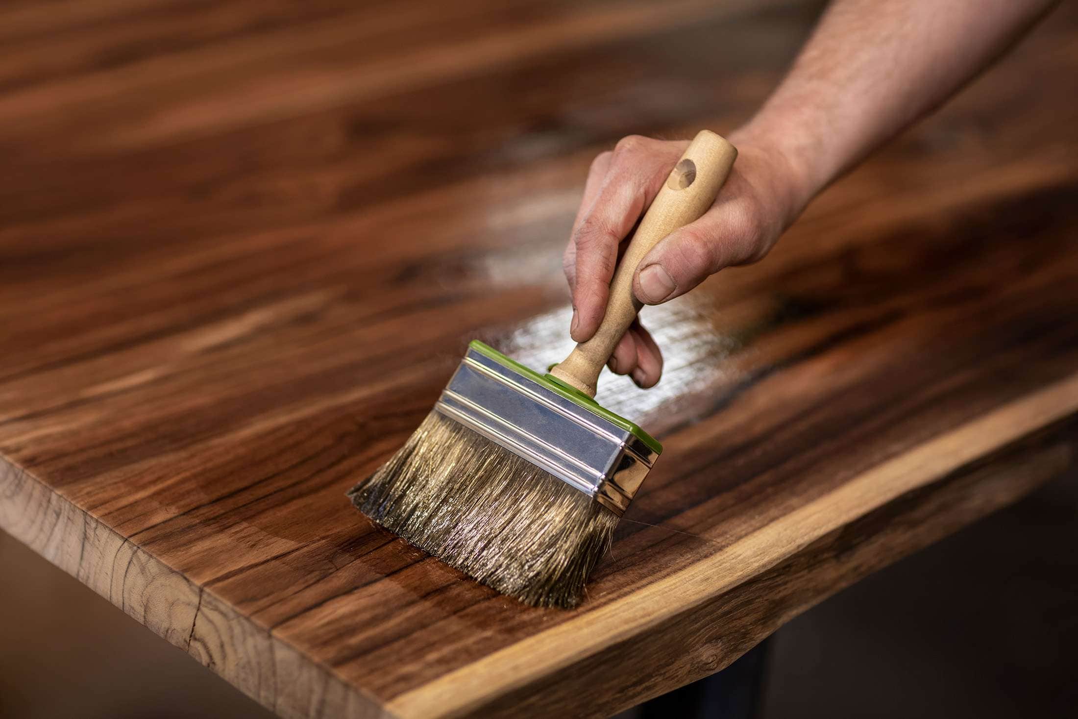 A hand is applying a clear finish to a wooden table using a large brush. The wood grain is visible, and the surface appears glossy from the finish, showcasing what skilled interior painting in Larimer County, CO can achieve.