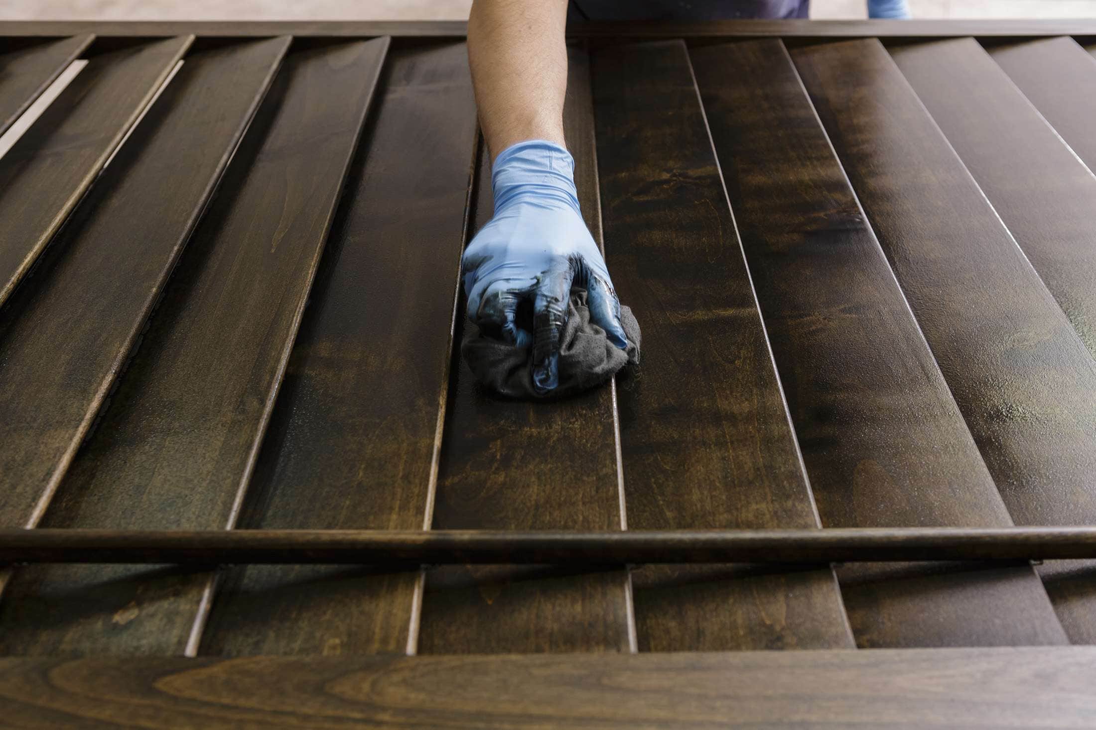 In Larimer County, CO, a person wearing blue gloves carefully applies stain to a wooden surface with a cloth. The close-up image highlights the intricate wood grain and the sheen from the finish, showcasing expert craftsmanship akin to top-tier painting services in Colorado.