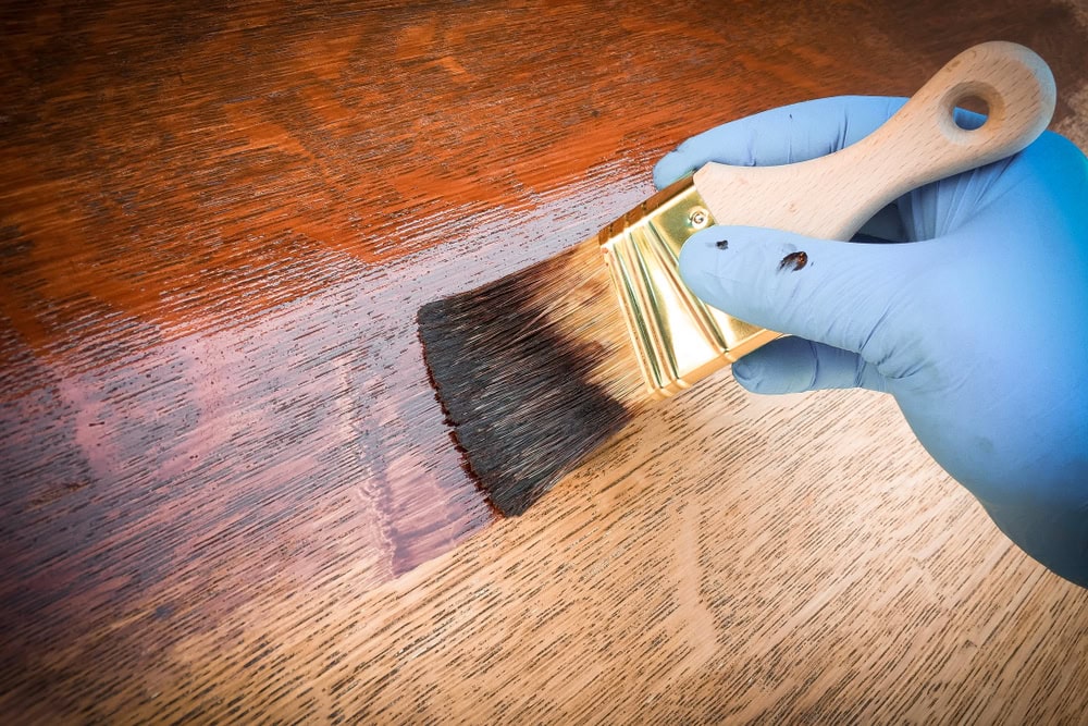 A gloved hand uses a paintbrush to apply dark wood stain to a light wooden surface, showing the contrast between the stained and unstained wood.