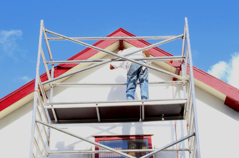 A person stands on scaffolding, performing residential exterior painting on the upper trim of a white house with a red roof, under a clear blue sky.