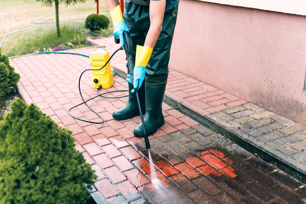 A person wearing gloves, boots, and protective clothing uses a pressure washer to clean a red brick walkway outside, with visible contrast between cleaned and dirty areas.