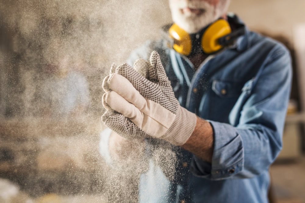 A man wearing work gloves, a denim shirt, and yellow ear protectors claps his hands, creating a cloud of dust in a workshop setting. His face is partially out of focus.