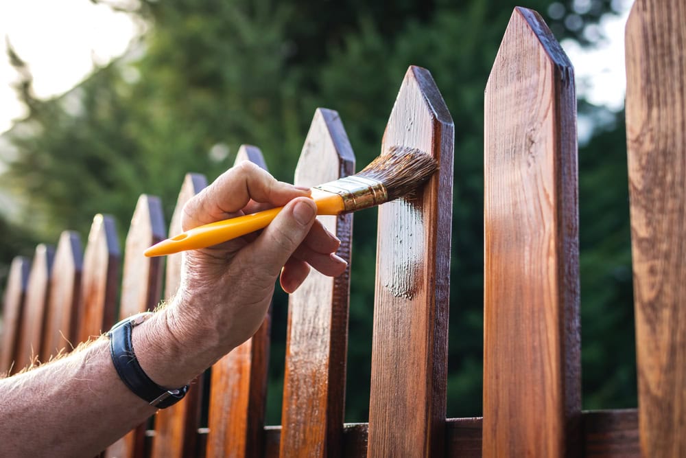 A person’s hand holds a yellow-handled paintbrush, applying varnish or paint to a wooden fence outdoors, with green trees blurred in the background.