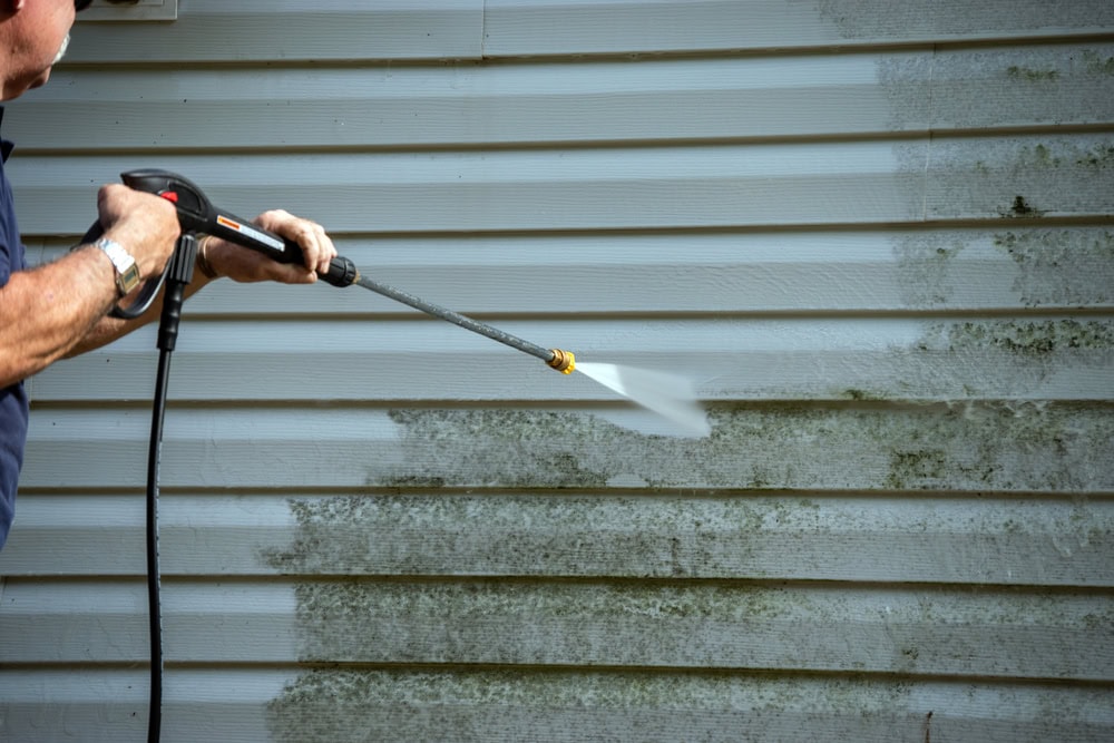 A person uses a pressure washer to clean mold and dirt off the siding of a house, revealing a much cleaner surface compared to the dirty, stained sections.