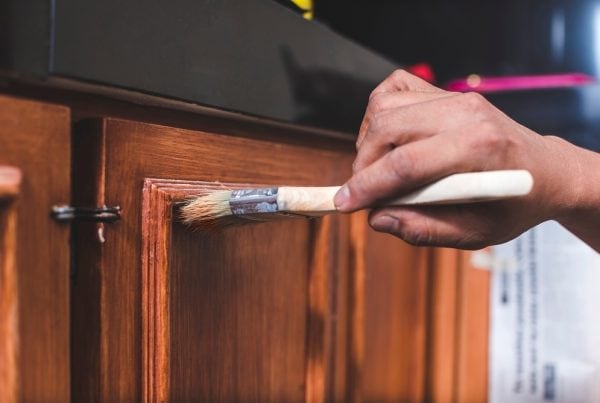 A close-up of a person painting a wooden cabinet door with a brush, applying a fresh coat of finish or paint. The hand and brush are in focus, highlighting the smooth strokes on the wood surface.