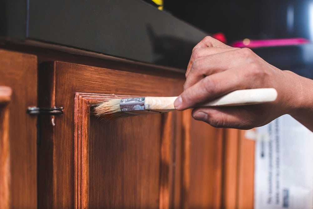 A close-up of a person painting a wooden cabinet door with a brush, applying a fresh coat of finish or paint. The hand and brush are in focus, highlighting the smooth strokes on the wood surface.