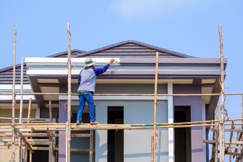 A worker standing on scaffolding paints the exterior trim of a modern house under construction, with a clear blue sky in the background.