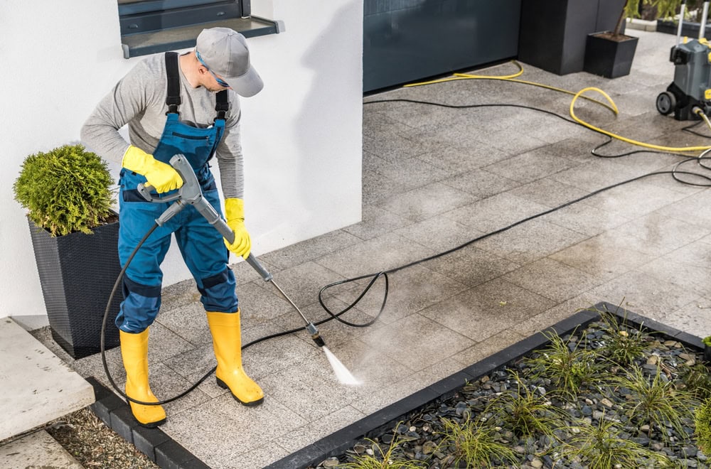 A person in yellow boots, gloves, and a gray cap uses a pressure washer to clean a tiled outdoor area next to a garden bed and a potted plant. Hoses are visible on the ground.