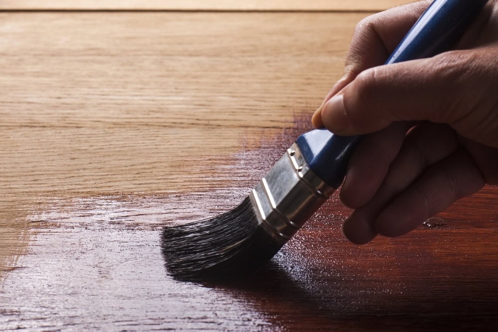 A hand holds a paintbrush, applying a dark wood stain or varnish to a wooden surface, highlighting the contrast between the treated and untreated sections of the wood.