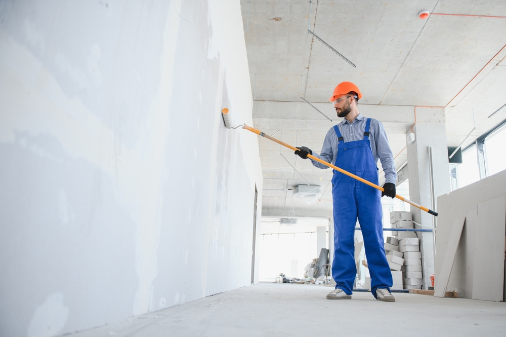 A construction worker in a hard hat, gloves, and blue overalls uses a paint roller with an extended handle to paint a large wall—part of a commercial exterior painting project on the construction site.