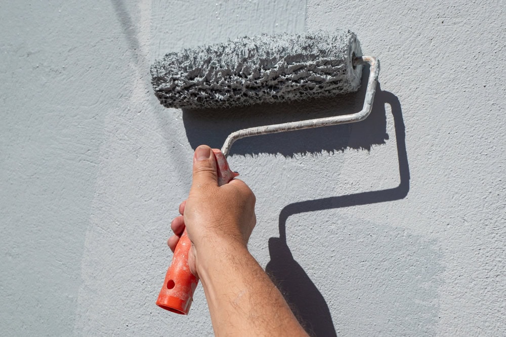 A person’s hand holding an orange-handled paint roller applies light gray paint to a wall, with the roller covered in wet paint and casting a shadow on the surface.