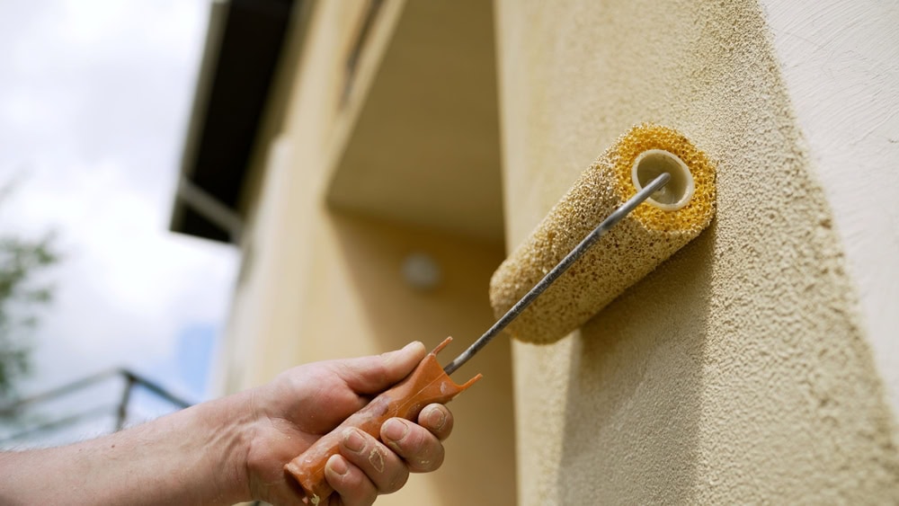 A hand holding a paint roller applies light beige paint to the exterior wall of a building on a sunny day.