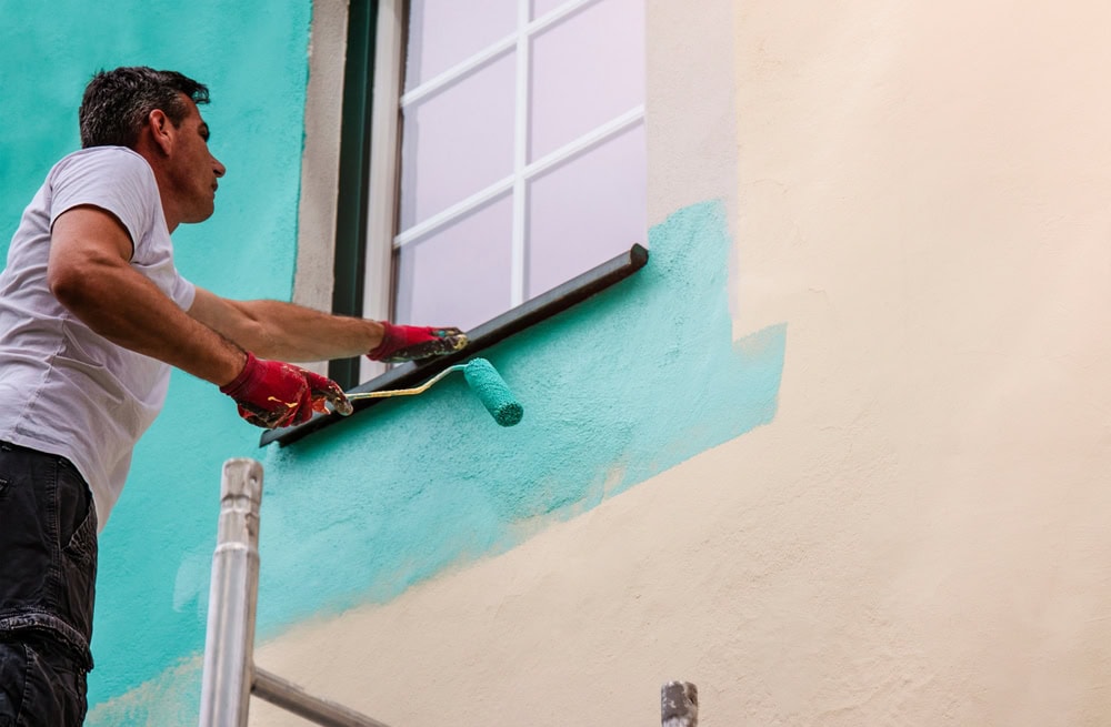 A man standing on a scaffold paints the exterior wall of a building turquoise using a paint roller, wearing red gloves and a white t-shirt, near a window with white frames.