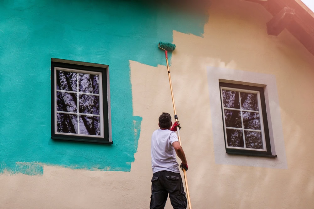 A person using a long-handled roller paints the exterior wall of a house from beige to teal, reaching above two windows.