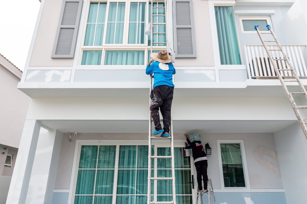 Two people are doing residential exterior painting, working on ladders near upper and lower windows. Both wear hats and sun-protective clothing as they paint the house’s exterior under the sun.