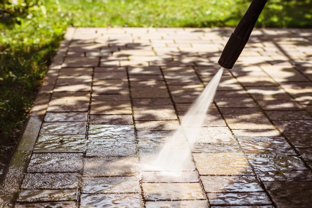 A pressure washer sprays water onto a paved patio, cleaning the surface. Sunlight filters through tree branches, creating dappled shadows on the wet and dry sections of the paving stones. Grass is visible in the background.