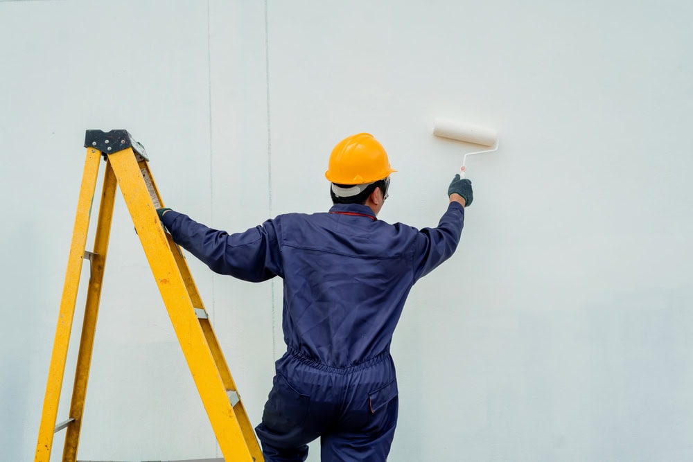 A worker in a blue jumpsuit and yellow hard hat uses a paint roller to paint a white wall, standing next to a yellow ladder.