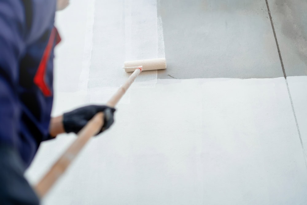 A person wearing a dark jacket and black gloves uses a paint roller on a long handle to paint a wall white, highlighting the precision often seen in commercial exterior painting. The photo is taken from above, focusing on the painting process.