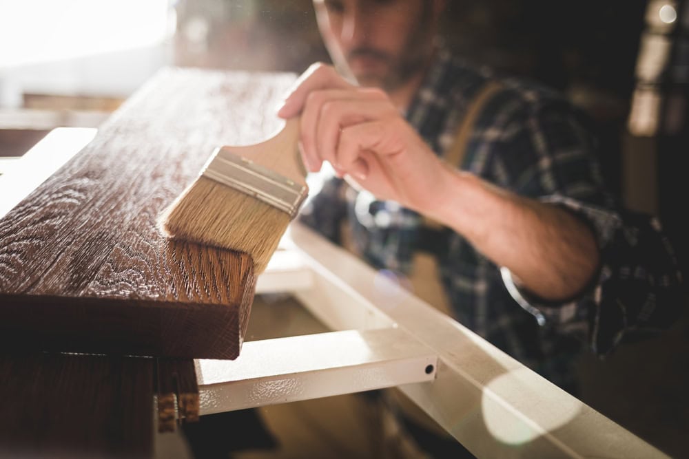 A person wearing a plaid shirt is applying varnish or wood stain to a wooden plank with a wide brush, working carefully in a well-lit workshop.