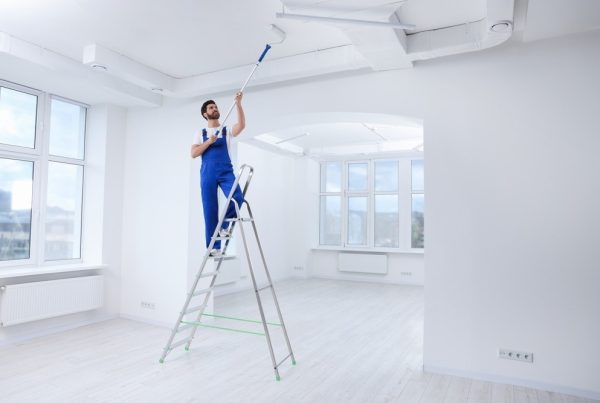A person in blue overalls stands on a ladder, painting a white ceiling with a roller in a bright, empty room with large windows and white walls.