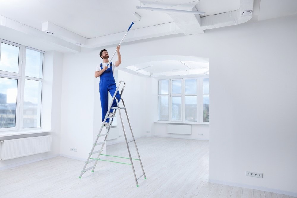 A person in blue overalls stands on a ladder, painting a white ceiling with a roller in a bright, empty room with large windows and white walls.