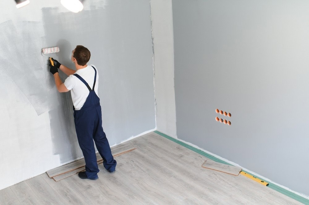 A person wearing navy overalls and black gloves paints a gray wall with a roller in a room with light wood flooring. Some wooden planks and tools are on the floor nearby.
