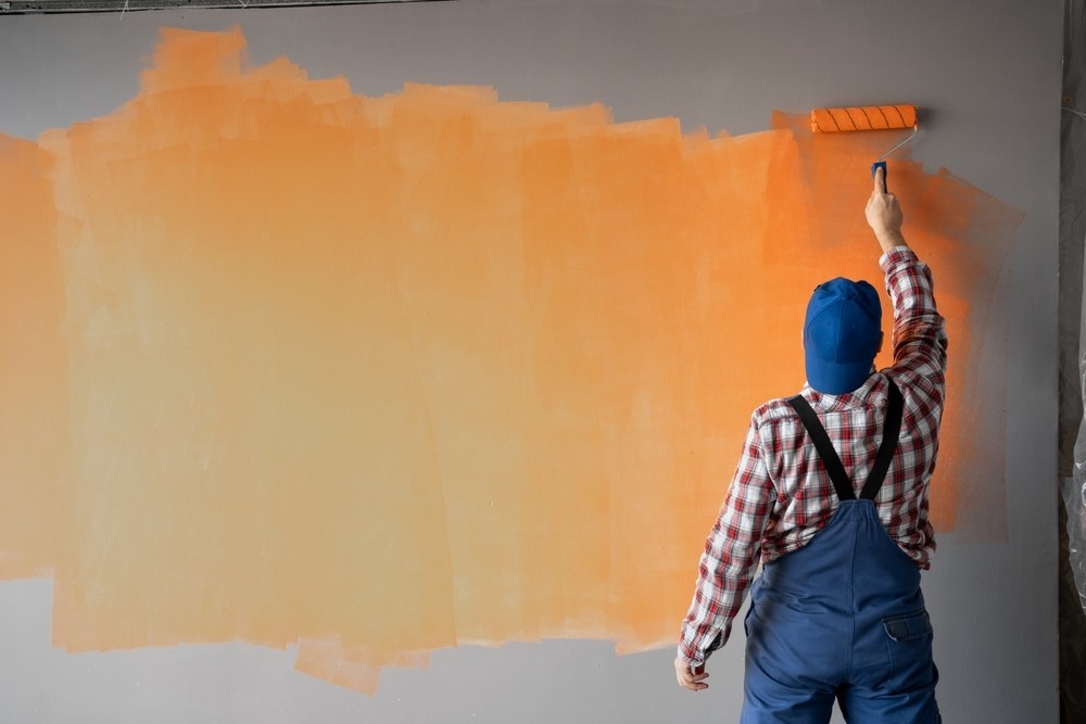 A person wearing blue overalls and a checked shirt paints a gray wall orange with a paint roller, standing with their back to the camera.