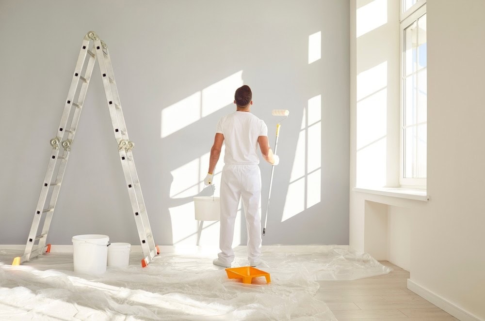 A person in white clothes stands in a bright room, painting a gray wall with a roller. A ladder, paint buckets, and a tray are on a plastic-covered floor near large sunlit windows.