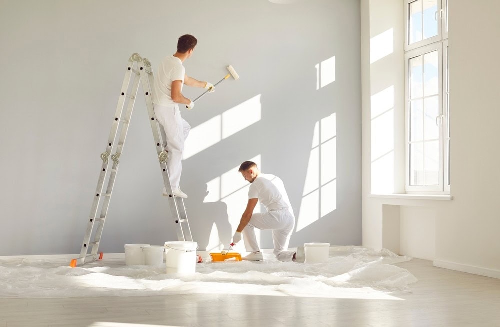 Two people dressed in white work clothes paint a light gray wall in a bright room. One person stands on a ladder using a roller, while the other crouches on the floor by paint buckets and a tray. Sunlight streams through large windows.