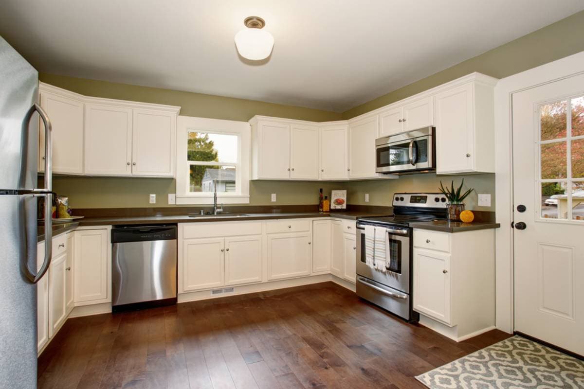 Modern kitchen with white cabinets, stainless steel appliances, dark countertops, and wood flooring. A window above the sink lets in natural light, and a door with glass panels opens to the outside.