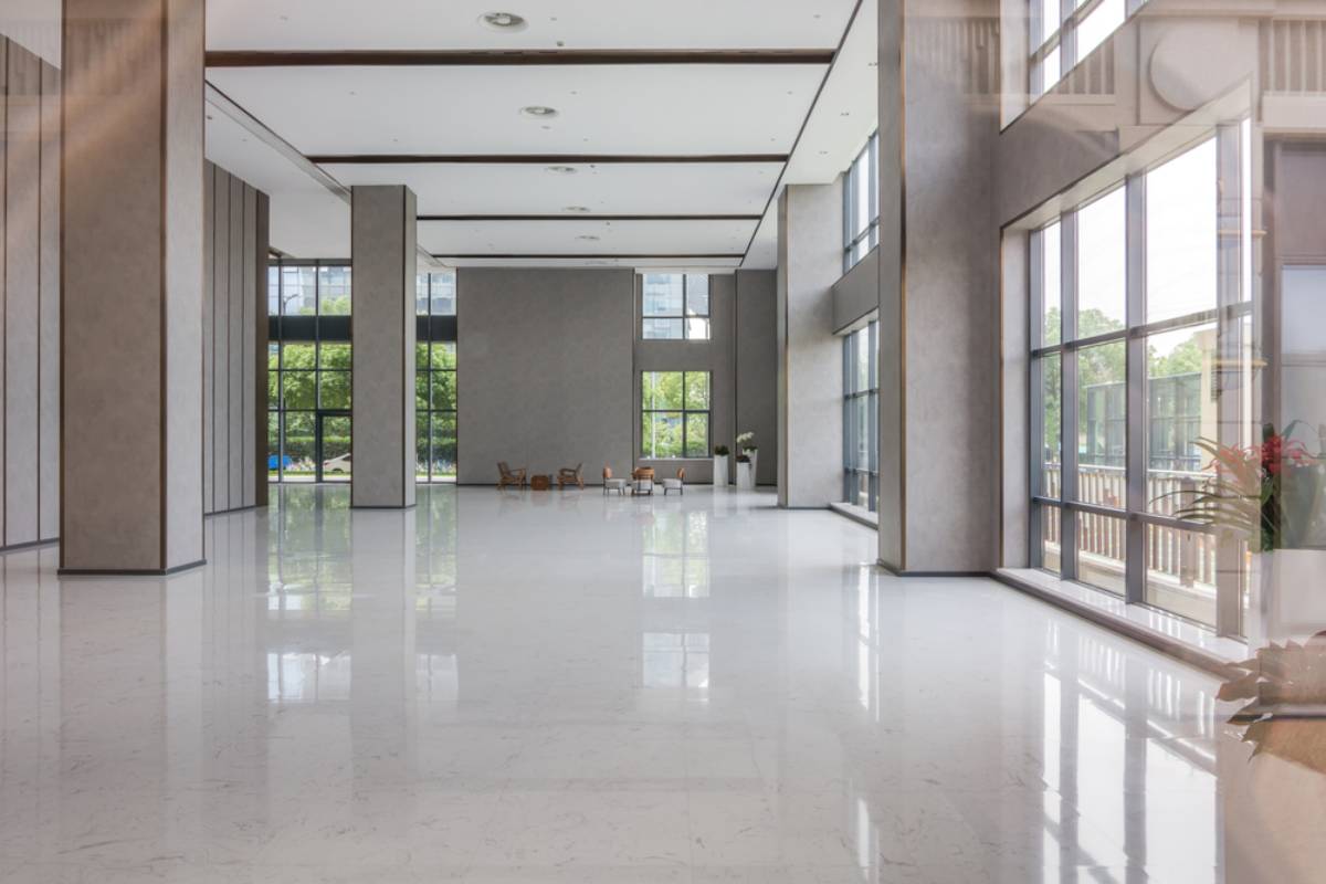 A spacious, modern lobby with high ceilings, large windows letting in natural light, polished white floors, a few chairs and tables in the distance, and potted plants near the windows.