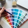 A hand holding a pen points to a color swatch fan deck displaying various shades of blue, with a laptop, paintbrush, and notebook visible on a desk in the background.
