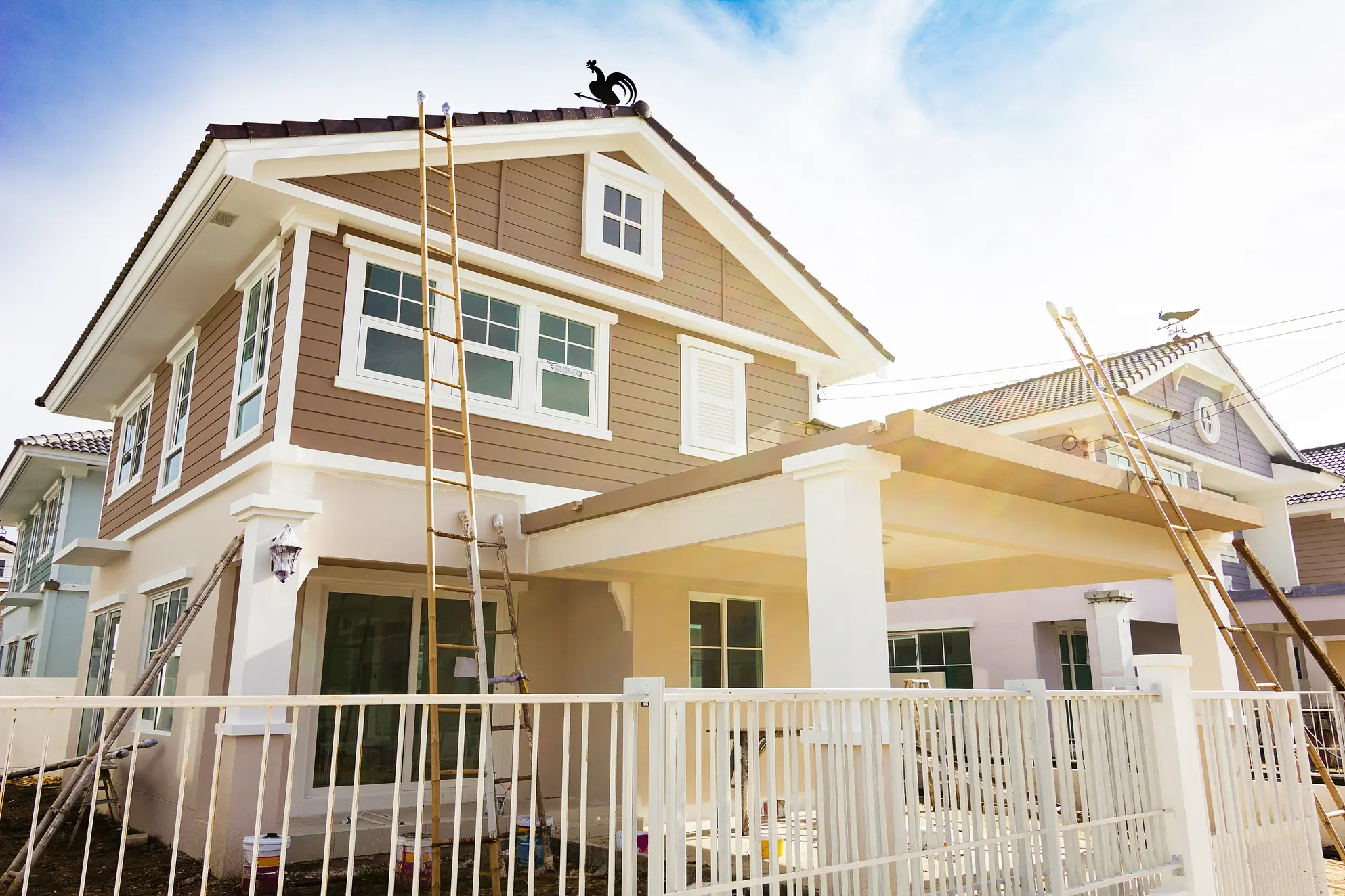 Two-story beige and white house with ladders set up for exterior painting, featuring large windows, a tiled roof, and a fenced yard under a bright blue sky in Larimer County, Colorado.