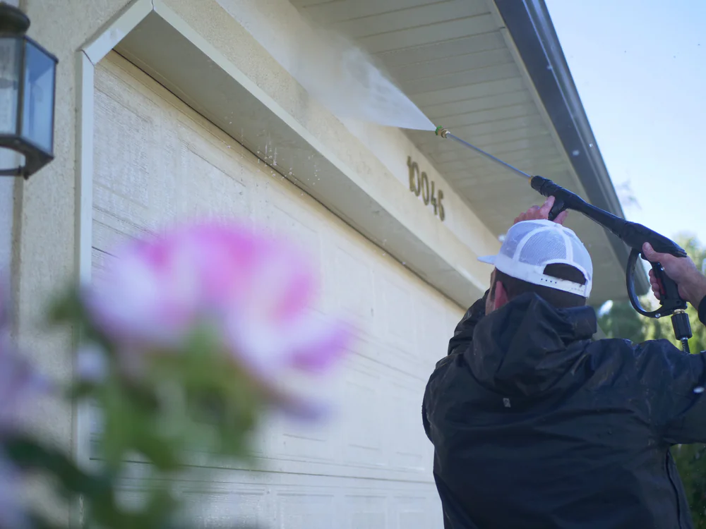 A person in a black jacket and white cap uses a pressure washer to clean the upper section of a garage door at a home in Larimer County, Colorado, with a pink flower blurred in the foreground.
